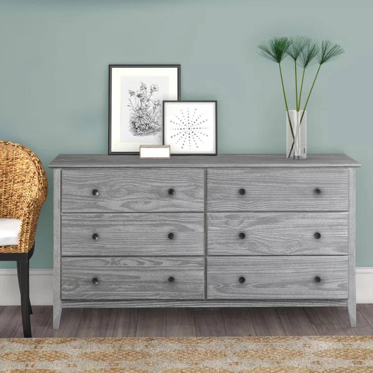 Gray weathered wood six-drawer dresser styled in modern bedroom with wicker chair, framed artwork, and decorative vase with dried branches